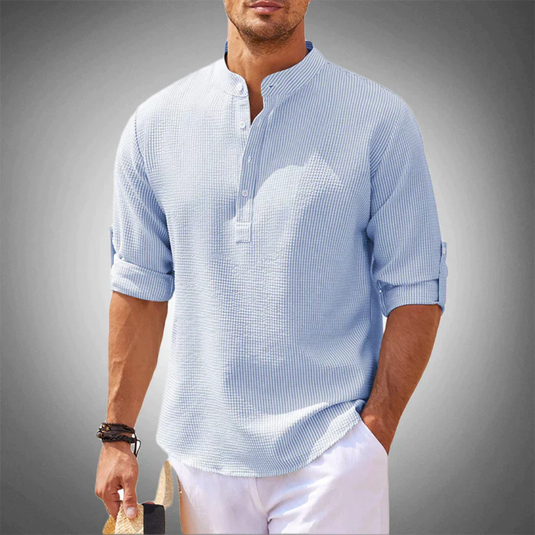 Mann in hellblauem Leinenhemd mit Stehkragen und hochgekrempelten Ärmeln, hält Strohhut. Sommermode, lässiger Stil, Herrenbekleidung.