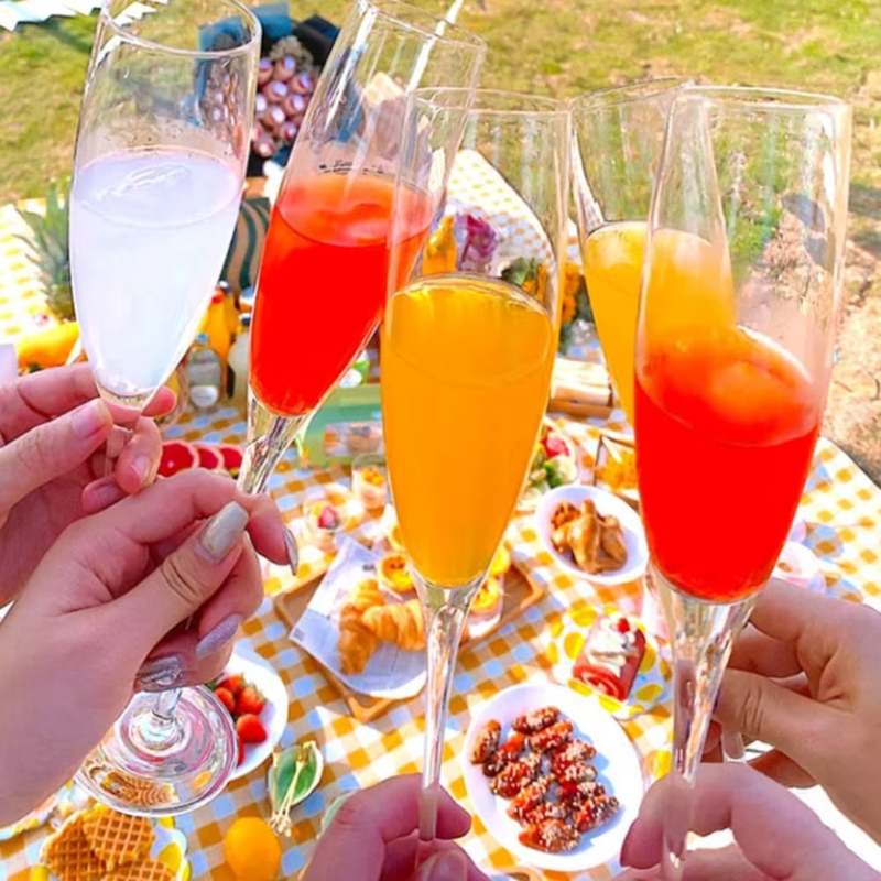 Laden Sie das Bild in Galerie -Viewer, Hände stoßen mit Sektgläsern an, gefüllt mit bunten Cocktails, auf einem Picknick im Freien. Sommer, Getränke, Feier, Picknick, Erfrischung.

