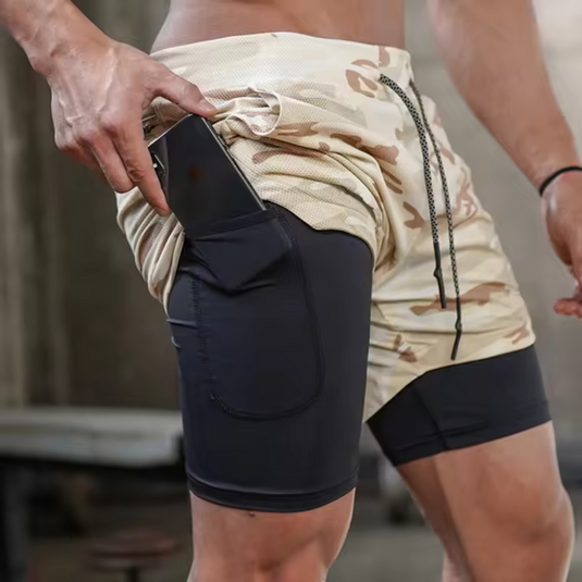 Mann in Camouflage-Shorts mit integriertem Handyfach, Fitnessstudio-Hintergrund, Sportbekleidung, praktisches Design für Training und Outdoor-Aktivitäten.