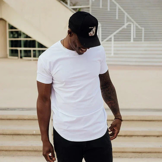Ein Mann in weißem T-Shirt und schwarzer Kappe steht lächelnd vor einer Treppe. Lässige Mode, urbaner Stil, Sommeroutfit.
