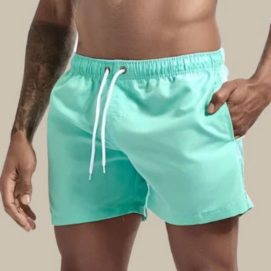 Mann in türkisfarbener Badehose mit Kordelzug, tätowierter Arm, neutraler Hintergrund. Sommermode, Herrenbadebekleidung, Strandoutfit.