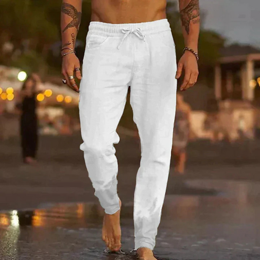 Mann in weißen Leinenhosen am Strand bei Sonnenuntergang, barfuß im Wasser. Sommermode, Freizeitkleidung, Strandspaziergang.