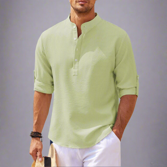 Mann in hellgrünem Leinenhemd mit Stehkragen und hochgekrempelten Ärmeln, hält Strohhut. Sommermode, Freizeitlook, Herrenbekleidung.