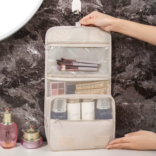 Beige hängender Kulturbeutel mit mehreren Fächern, gefüllt mit Kosmetikartikeln und Pinseln, auf Marmorhintergrund. Ideal für Reisen und Badezimmerorganisation.