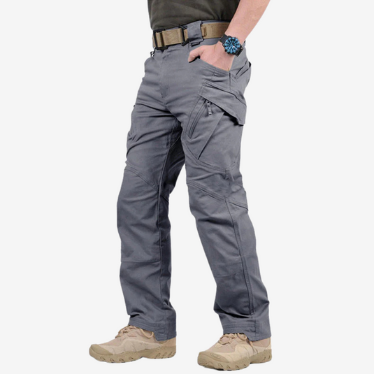 Mann trägt graue taktische Hose mit mehreren Taschen, beiger Gürtel und beige Stiefel. Outdoor-Mode, funktionale Kleidung, robuste Materialien.