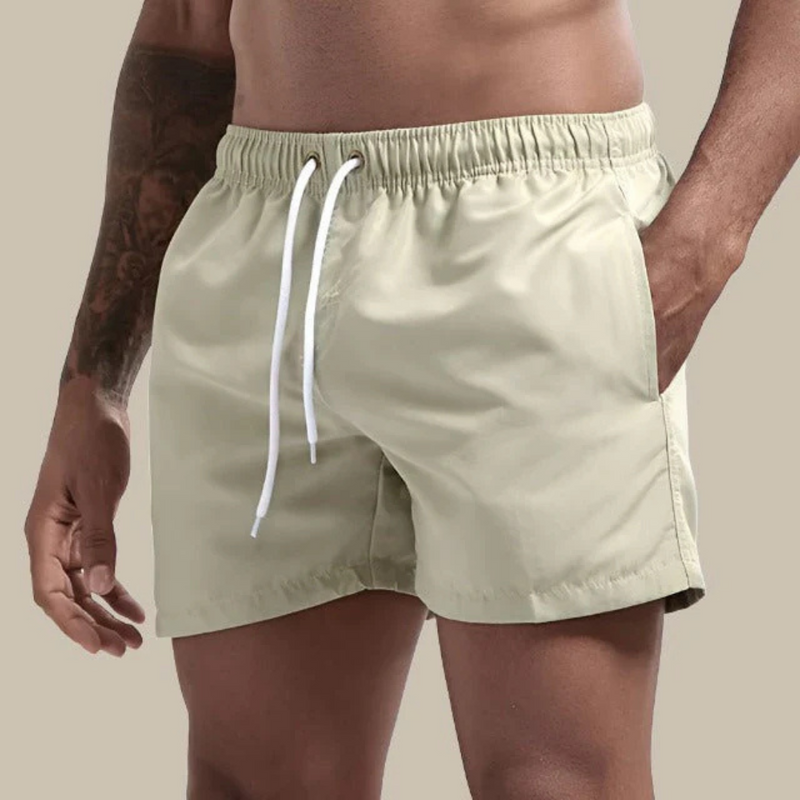 Laden Sie das Bild in Galerie -Viewer, Mann in beigen Badeshorts mit weißem Kordelzug, tätowierter Arm, neutraler Hintergrund. Sommermode, Herrenbadehose, Strandbekleidung.
