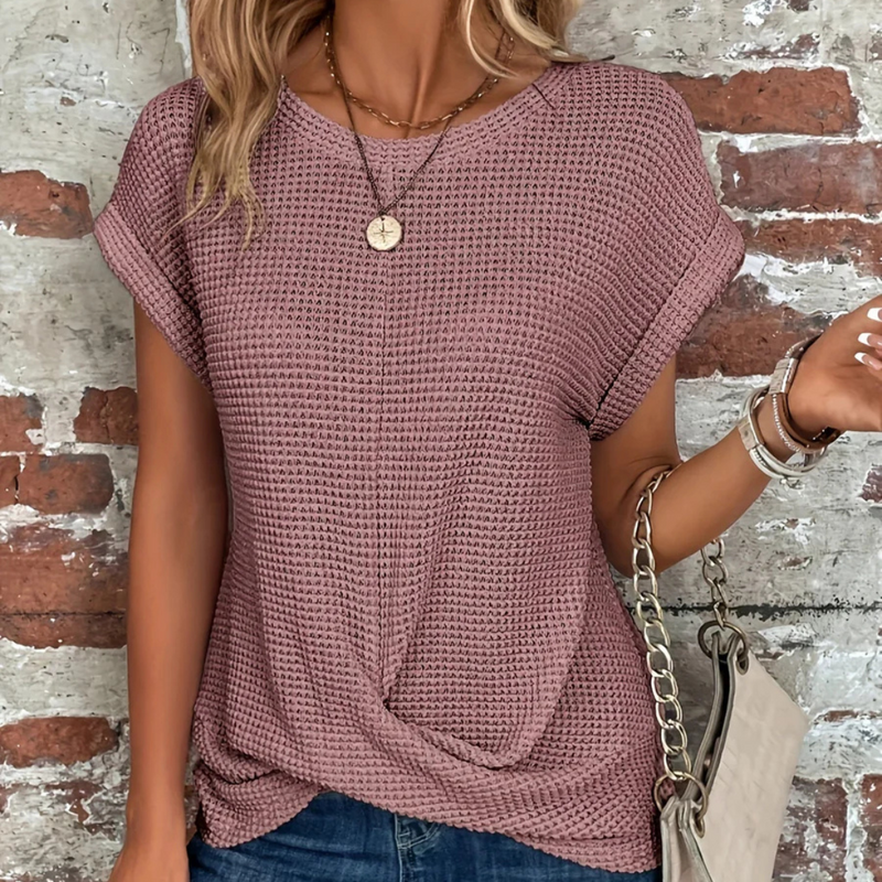Laden Sie das Bild in Galerie -Viewer, Frau in lila Strickshirt vor Ziegelwand, trägt Halskette und Armbänder, hält beigefarbene Handtasche. Modetrend, lässiger Look, Damenmode.
