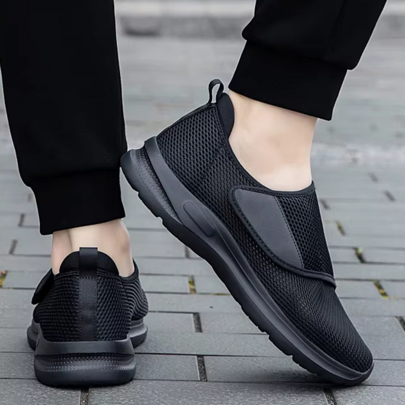 Laden Sie das Bild in Galerie -Viewer, Schwarze Herren-Sneaker mit Klettverschluss, atmungsaktivem Mesh-Obermaterial und rutschfester Sohle auf grauem Pflasterweg.
