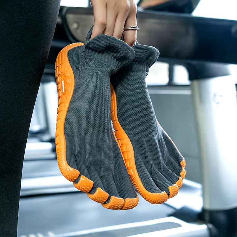 Laden Sie das Bild in Galerie -Viewer, Dunkelgraue Barfußschuhe mit orangefarbener Sohle, gehalten von einer Hand im Fitnessstudio. Ideal für minimalistisches Training und natürliche Fußbewegung.
