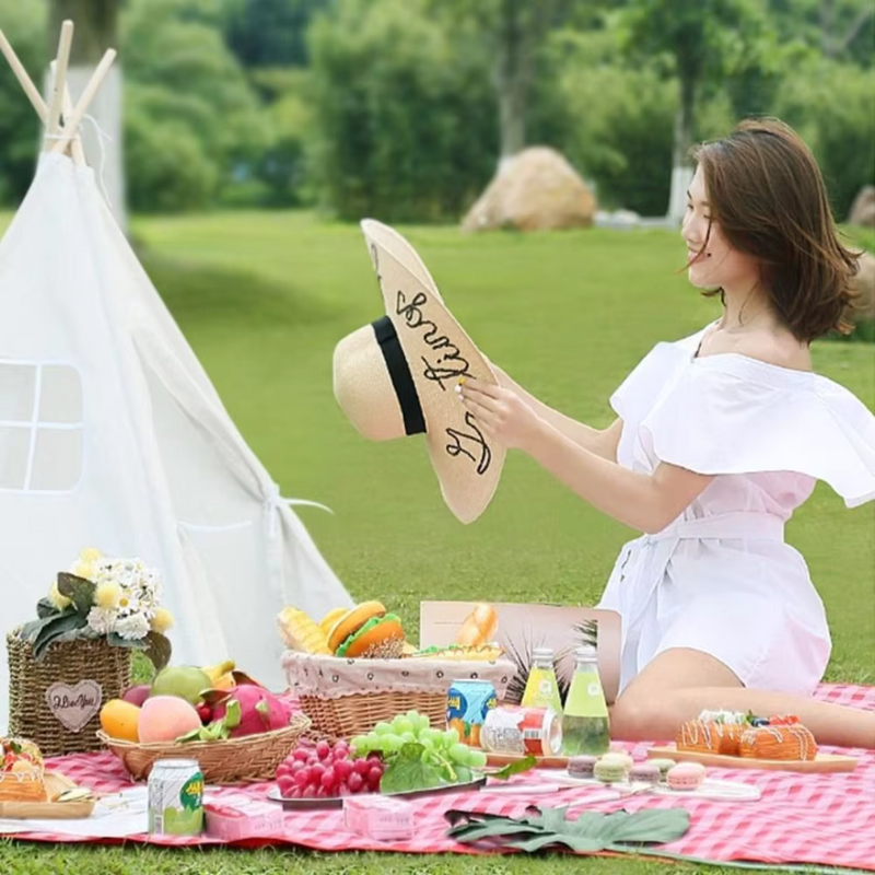 Laden Sie das Bild in Galerie -Viewer, Frau im weißen Kleid bei Picknick auf Wiese, hält Sonnenhut. Vor ihr Obstkorb, Getränke, Zelt. Sommer, Natur, Freizeit, Entspannung im Freien.
