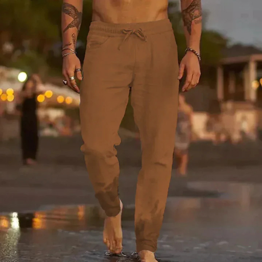 Ein Mann in brauner Jogginghose geht barfuß am Strand entlang. Abendstimmung, verschwommener Hintergrund mit Lichtern und Menschen.