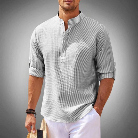Mann in grauem Leinenhemd mit Stehkragen und hochgekrempelten Ärmeln, hält Strohhut. Sommermode, lässiger Stil, Herrenbekleidung.