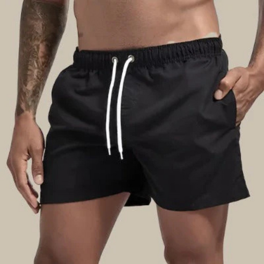 Mann in schwarzen Badeshorts mit Kordelzug, tätowierter Arm, beige Hintergrund. Herrenmode, Sommerbekleidung, Strandoutfit.