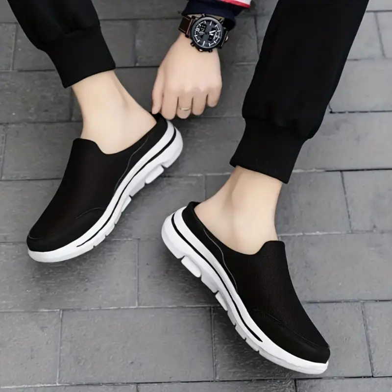 Laden Sie das Bild in Galerie -Viewer, Schwarze Herren-Sneaker-Slipper mit weißer Sohle auf grauem Pflaster, getragen von Person in schwarzer Hose. Bequeme, modische Freizeitschuhe.
