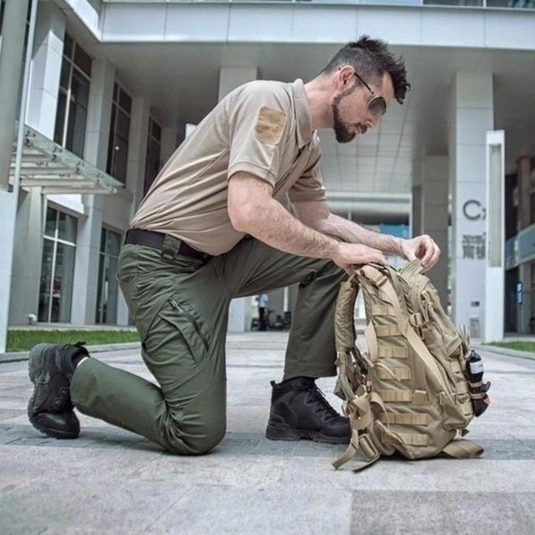 Mann in taktischer Kleidung kniet vor modernem Gebäude und überprüft beigen Rucksack. Outdoor-Ausrüstung, taktische Mode, urbane Umgebung.