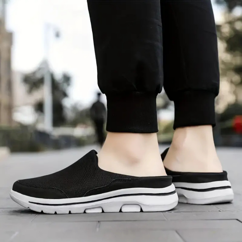 Laden Sie das Bild in Galerie -Viewer, Schwarze Slip-On-Sneaker mit weißer Sohle auf Gehweg, getragen von Person in schwarzer Hose. Bequeme Freizeitschuhe, urbaner Stil.
