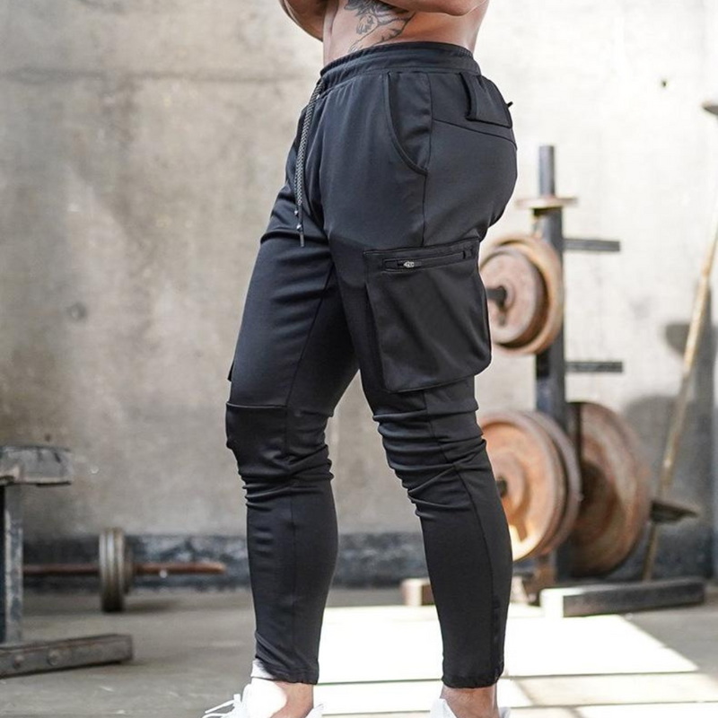 Laden Sie das Bild in Galerie -Viewer, Mann in schwarzer Jogginghose mit Seitentaschen im Fitnessstudio, Hanteln im Hintergrund. Sportbekleidung, Fitnessmode, Herren-Trainingshose.
