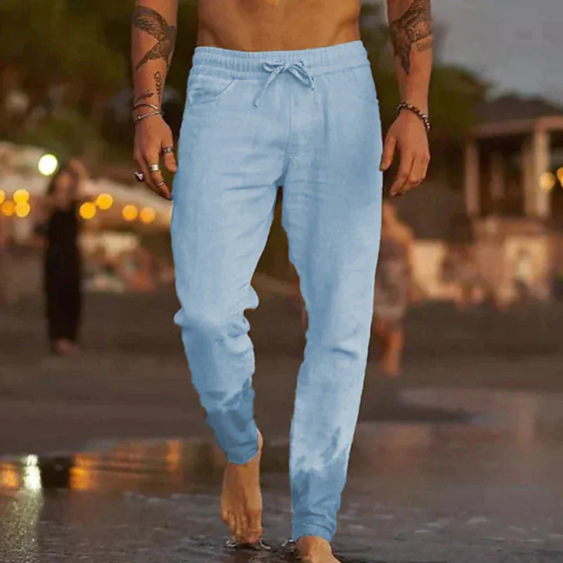 Laden Sie das Bild in Galerie -Viewer, Mann in hellblauer Jogginghose am Strand bei Sonnenuntergang, barfuß im Sand. Freizeitmode, Sommerkleidung, Strandspaziergang.
