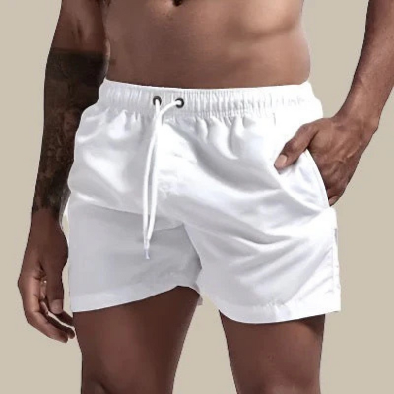 Laden Sie das Bild in Galerie -Viewer, Mann in weißen Badeshorts, muskulöser Oberkörper, tätowierter Arm, beige Hintergrund. Sommermode, Herrenbadehose, Strandbekleidung.
