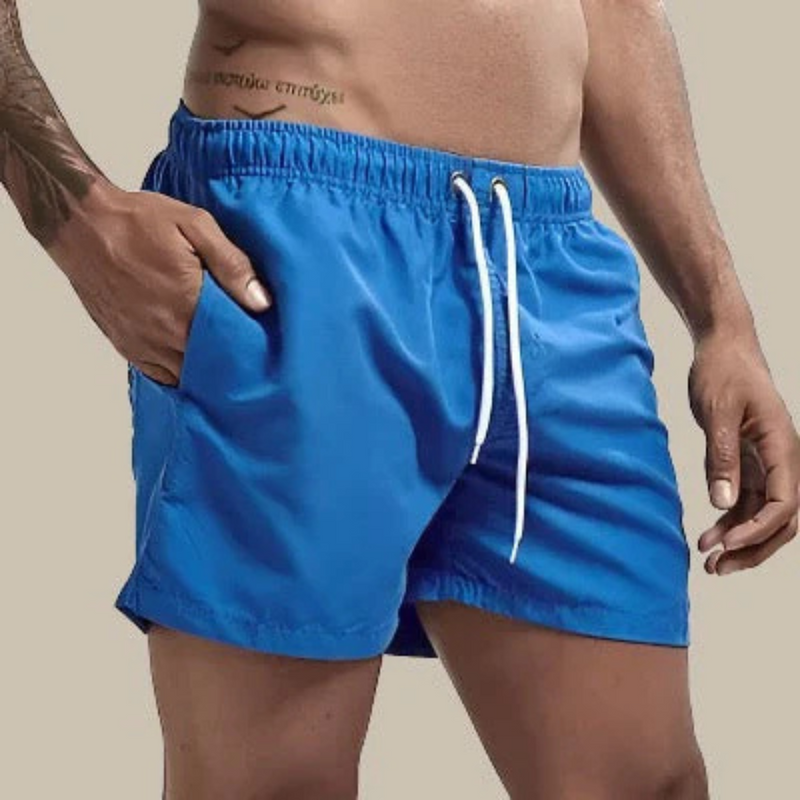Laden Sie das Bild in Galerie -Viewer, Mann in blauer Badehose mit Kordelzug, tätowierter Arm, beige Hintergrund. Sommermode, Herrenbademode, Strandbekleidung.

