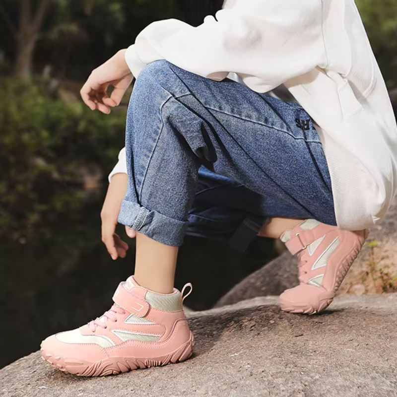 Laden Sie das Bild in Galerie -Viewer, Frau in blauer Jeans und weißem Pullover trägt rosa Wanderschuhe mit Klettverschluss, sitzt auf einem Felsen im Freien. Outdoor-Mode, bequeme Schuhe.
