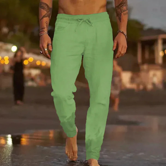 Mann in grüner Leinenhose am Strand bei Sonnenuntergang, barfuß im Wasser. Sommermode, Freizeitkleidung, Strandspaziergang.